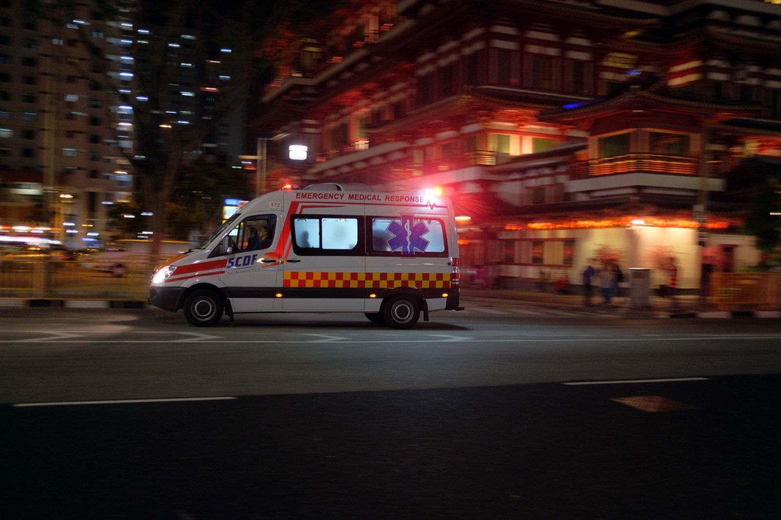 An SCDF emergency ambulance marked Emergency Medical Response speeds through Singapore's Chinatown at night, past the illuminated Buddha Tooth Relic Temple.
